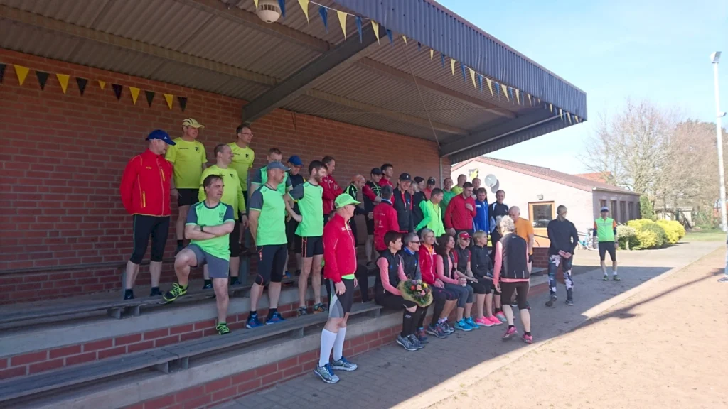 Gruppenfoto von Sportlern in Laufbekleidung auf einer Tribüne, umgeben von farbigen Wimpeln. Die Teilnehmer stehen und sitzen in verschiedenen Positionen, während sie sich auf ein Event vorbereiten.