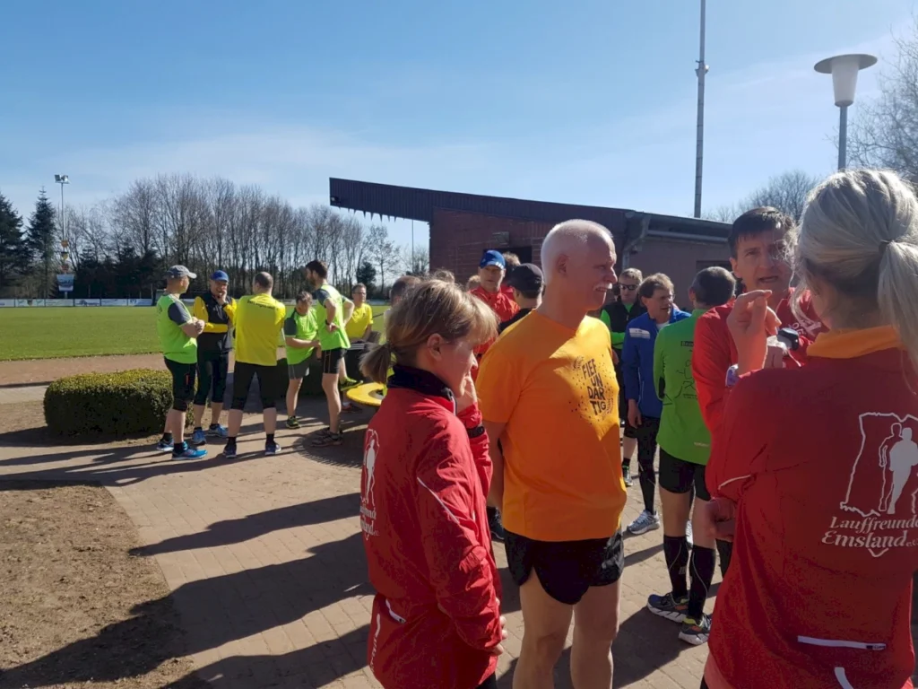 Gruppierung von Läufern in bunten Trikots, die sich vor einem Sportplatz unterhalten. Der sonnige Himmel und die frühlingshafte Atmosphäre schaffen eine motivierende Umgebung für sportliche Aktivitäten. Im Hintergrund sind weitere Teilnehmer und Sporteinrichtungen sichtbar.