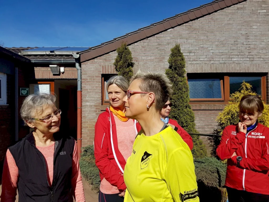 Gruppe von Frauen in Sportkleidung, die sich im Freien unterhalten, vor einem Gebäude mit Solarpanelen. Die Szene zeigt eine freundliche Atmosphäre und aktive Lebensweise.