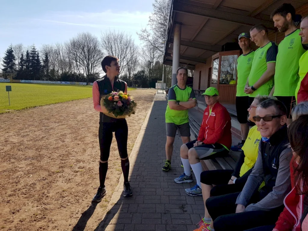 Eine Frau mit einem Blumenstrauß steht auf einem Sportplatz, während eine Gruppe von Läufern in leuchtend grünen T-Shirts auf einer Bank sitzt. Der Himmel ist klar und sonnig, und die Szene vermittelt eine freundliche, sportliche Atmosphäre.
