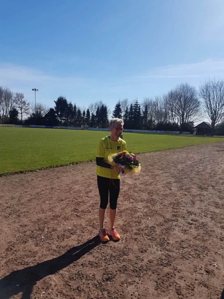 Frau in gelbem Sporttrikot steht auf einem Fußballplatz und hält einen Blumenstrauß in der Hand, umgeben von grünem Rasen und blauem Himmel.