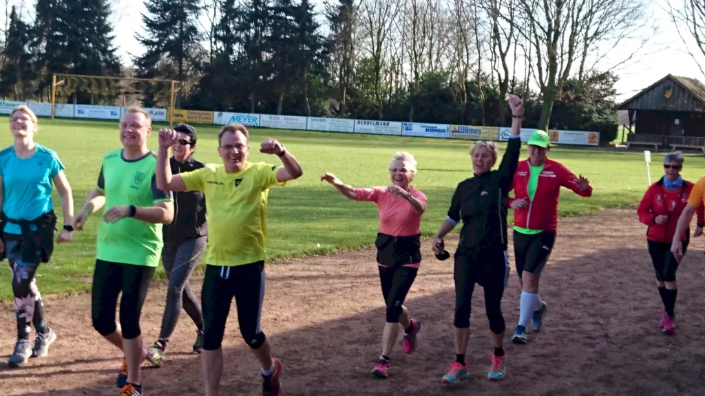 Eine Gruppe von Menschen in Sportkleidung läuft fröhlich auf einem Sportplatz, umgeben von Bäumen und einer grünen Wiese. Sie zeigen Freude und Begeisterung beim Laufen, was auf eine Gemeinschaftsveranstaltung oder ein Training hindeutet.