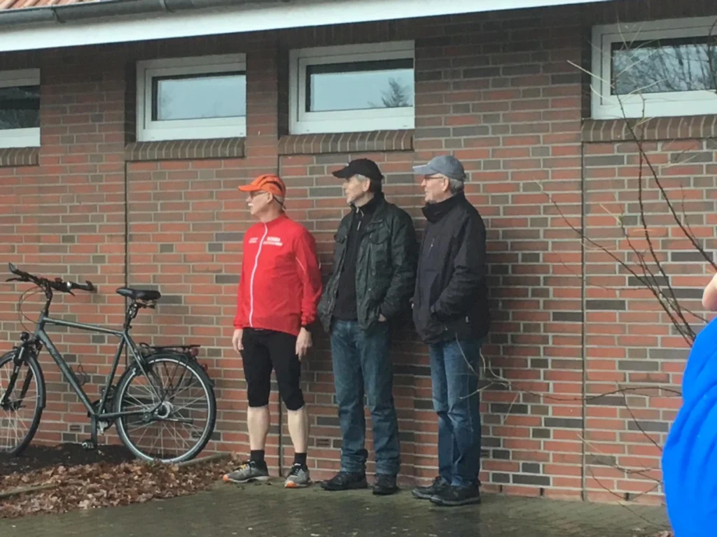 Drei Männer stehen an einer Ziegelwand, während ein Fahrrad neben ihnen lehnt. Der Mann links trägt eine rote Sportjacke und eine orange Kappe, während die anderen beiden in dunklerer Kleidung gekleidet sind. Die Szene spielt sich an einem bewölkten Tag ab, umgeben von herbstlichem Laub.