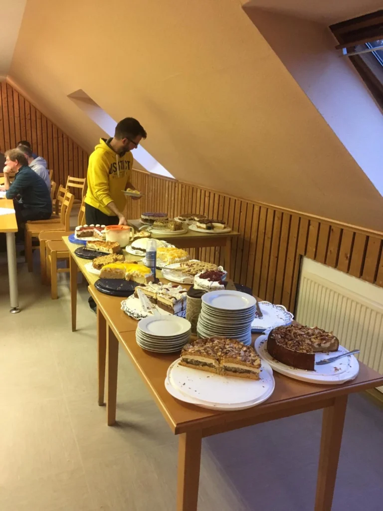 Ein Mann in einem gelben Sweatshirt steht an einem Tisch mit einer Auswahl an verschiedenen Kuchen und Torten, während im Hintergrund weitere Personen sitzen. Der Tisch ist mit Tellern und einer Vielzahl von Desserts gedeckt.