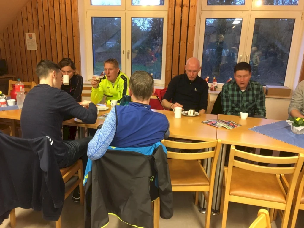 Gruppe von Männern und einer Frau beim geselligen Beisammensein in einem Raum mit Holzverkleidung. Sie sitzen an einem Tisch, trinken aus Tassen und essen Snacks. Auf dem Tisch sind Getränke und Zeitschriften sichtbar.