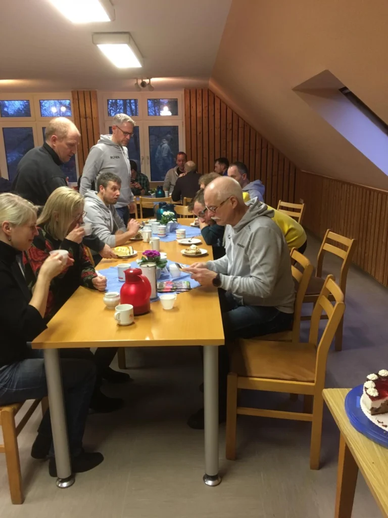 Eine Gruppe von Menschen sitzt an einem Tisch in einem gemütlichen Raum und genießt eine Mahlzeit. Auf dem Tisch stehen verschiedene Speisen und Getränke, darunter eine rote Teekanne und ein Kuchen. Die Atmosphäre ist freundlich und gesellig, mit Personen, die miteinander kommunizieren und das Essen teilen.