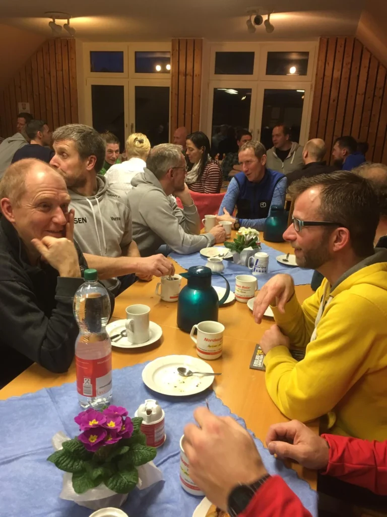 Gruppengespräch bei einem gemütlichen Abendessen in einem Holzraum, mit mehreren Personen an einem Tisch, der mit Tassen, Wasserflaschen und Blumen dekoriert ist. Die Atmosphäre ist entspannt und gesellig.
