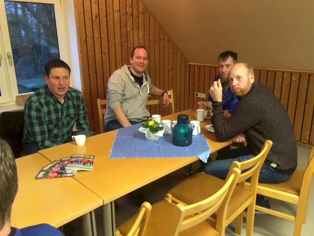 Gruppe von vier Männern, die an einem Tisch in einem gemütlichen Raum sitzen, mit Kaffeetassen und Snacks. Auf dem Tisch liegt eine blaue Tischdecke und einige Magazine. Holzvertäfelung im Hintergrund sorgt für eine warme Atmosphäre.