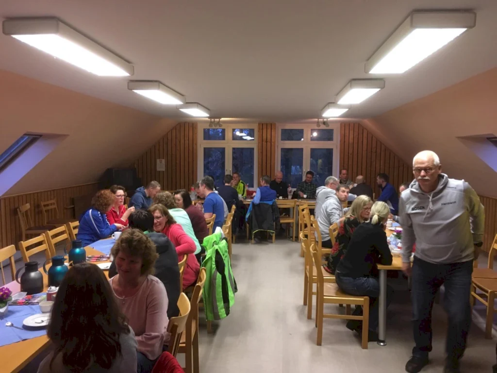 Gruppensitzung in einem gemütlichen Raum mit Holzverkleidung, wo Menschen an Tischen sitzen und sich unterhalten. Einige Teilnehmer genießen Snacks und Getränke, während andere aktiv an Gesprächen teilnehmen. Helle Deckenleuchten sorgen für eine einladende Atmosphäre.