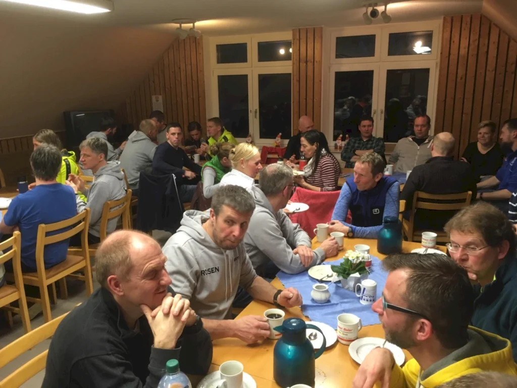 Gruppensitzung in einem gemütlichen Speisesaal mit mehreren Personen, die an Tischen sitzen, Essen teilen und Gespräche führen. Die Atmosphäre ist gesellig und entspannt, mit Tassen und Tellern auf den Tischen.