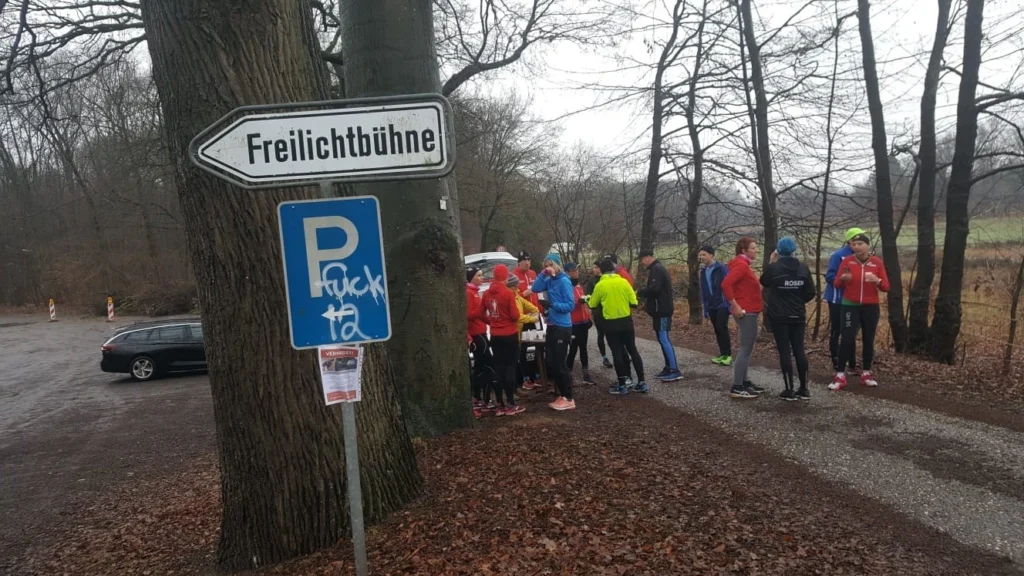Gruppe von Läufern in Sportkleidung versammelt sich an einem Parkplatz unter einem Schild mit der Aufschrift "Freilichtbühne". Im Hintergrund sind Bäume und eine regnerische Landschaft zu sehen.