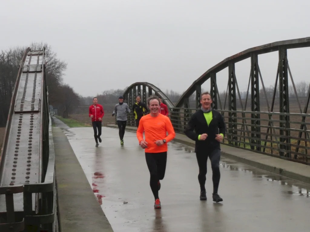 Gruppe von Läufern in Sportbekleidung joggt auf einer Brücke an einem bewölkten Tag. Die Strecke ist nass und spiegelt die Umgebung wider, während die Teilnehmer in verschiedenen Farben, darunter Orange und Rot, sichtbar sind.