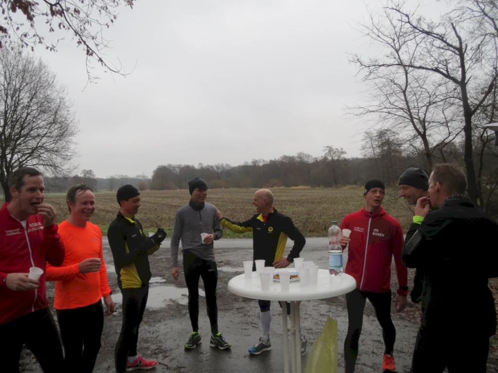 Gruppe von Läufern in sportlicher Kleidung, die an einem Tisch stehen und sich unterhalten, während sie ein Getränk genießen. Im Hintergrund eine herbstliche Landschaft mit Bäumen und einem bewölkten Himmel.