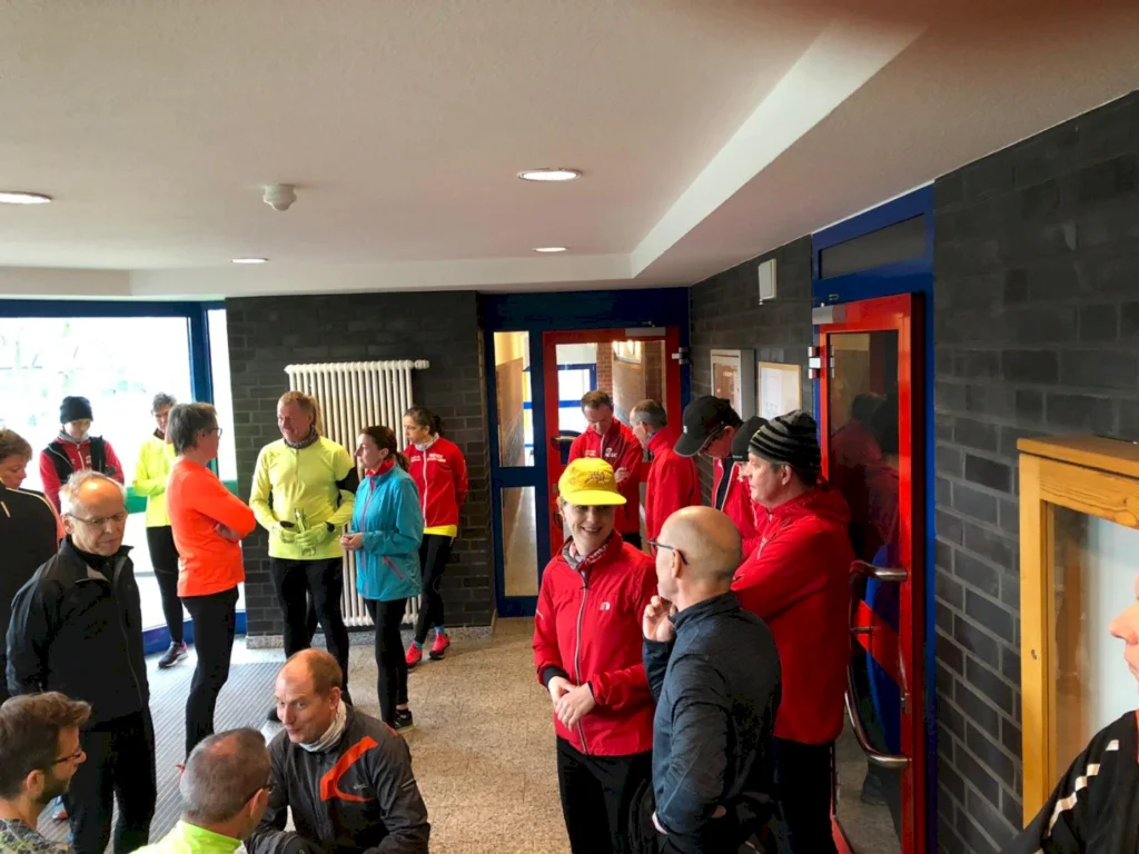 Gruppentreffen von Läufern in einem Sportgebäude, Mitglieder in bunten Laufoutfits stehen und unterhalten sich, während sie auf den Start eines Events warten.