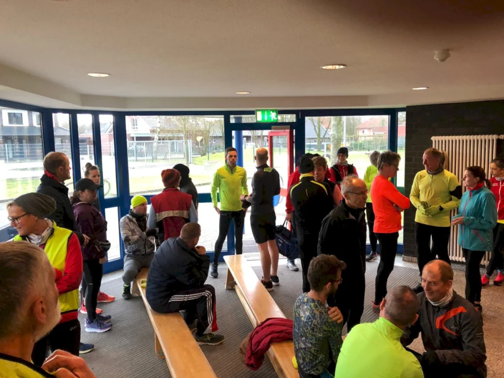 Gruppen von Läufern in sportlicher Kleidung stehen und unterhalten sich in einem hellen Foyer, während sie sich auf einen Lauf vorbereiten.