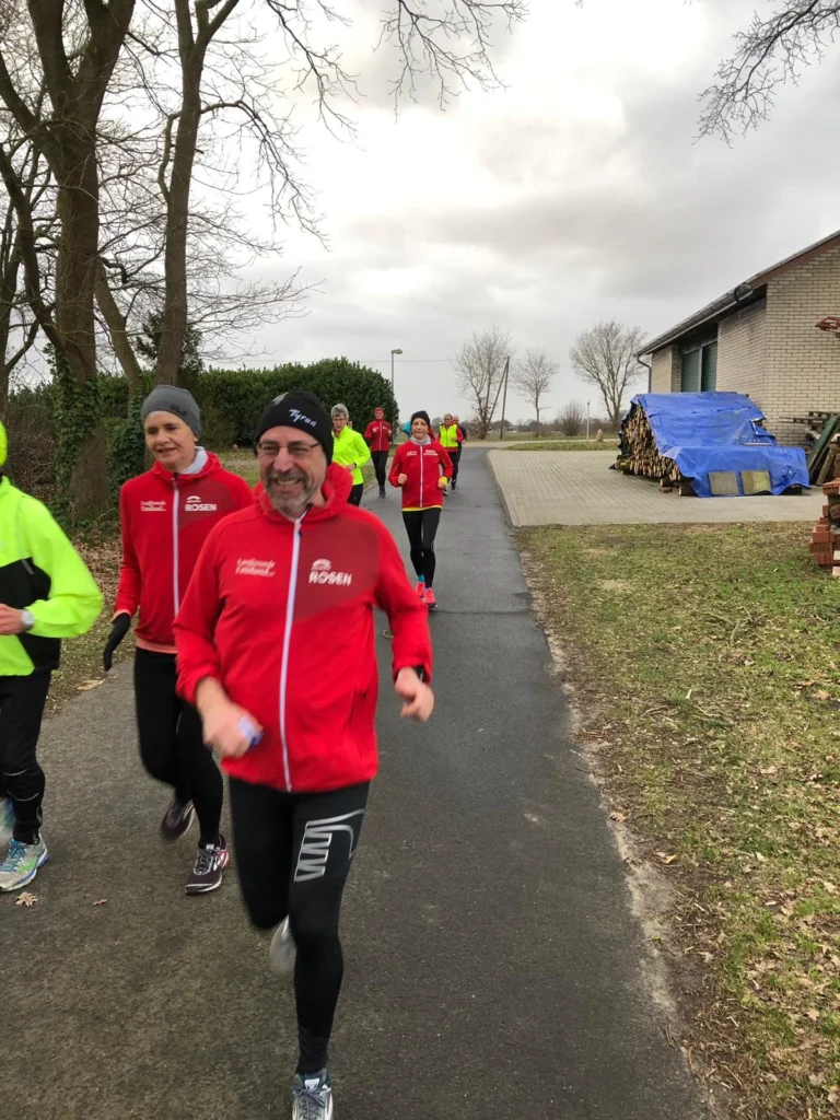 Läufer in roten Jacken joggen auf einem Weg, umgeben von Bäumen und einem Wohnhaus. Einige Teilnehmer tragen auch leuchtend gelbe Kleidung. Der Himmel ist bewölkt, und die Gruppe scheint sich aktiv zu unterhalten.
