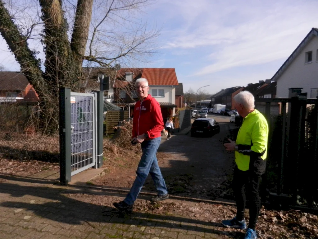 Zwei Männer beim Spaziergang in einer ruhigen Wohngegend, einer trägt eine rote Jacke und der andere ein leuchtend gelbes Shirt. Im Hintergrund sind Häuser und parkende Autos sichtbar, während Bäume und ein Zaun die Szene umrahmen.