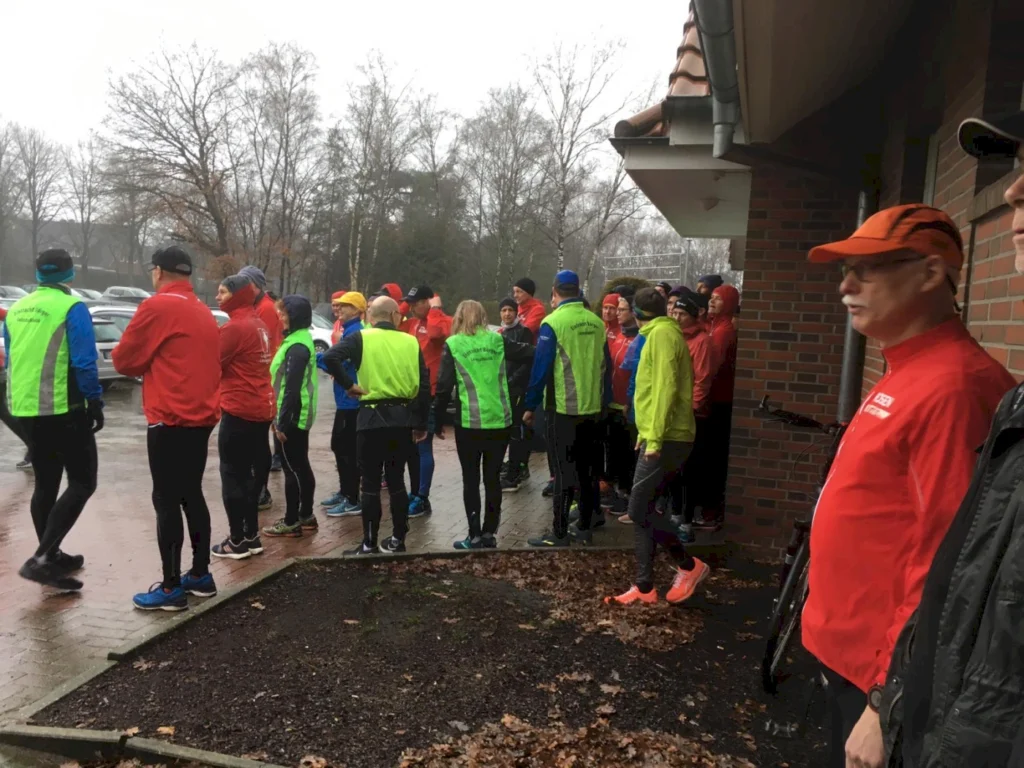 Gruppe von Läufern in Regenkleidung, versammelt vor einem Gebäude, während es regnet. Die Teilnehmer tragen leuchtend rote und grüne Jacken, bereit für ein gemeinsames Lauftraining.