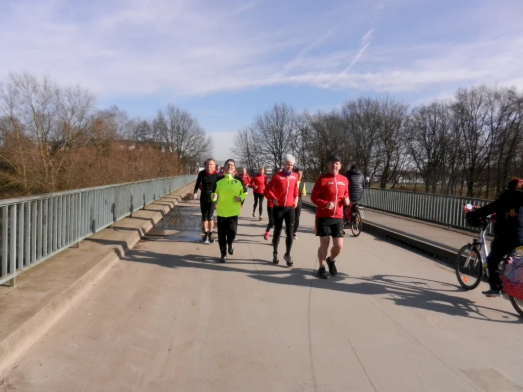 Gruppe von Läufern in roten Trikots, darunter ein Läufer in einem grünen Shirt, der über eine Brücke joggt. Im Hintergrund sind Fahrradfahrer und kahle Bäume zu sehen, bei klarem Himmel und sonnigem Wetter.