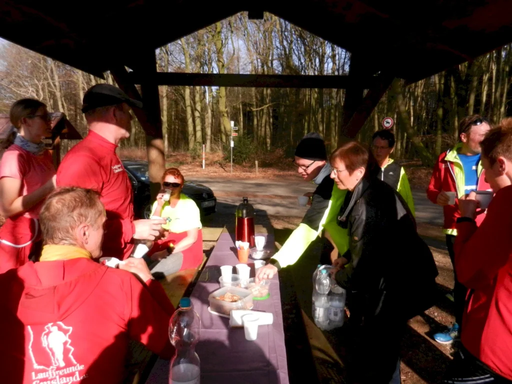 Gruppe von Läufern bei einer Pause im Wald, die Getränke und Snacks an einem Tisch genießen. Die Teilnehmer tragen Sportbekleidung in verschiedenen Farben und stehen unter einem Unterstand.