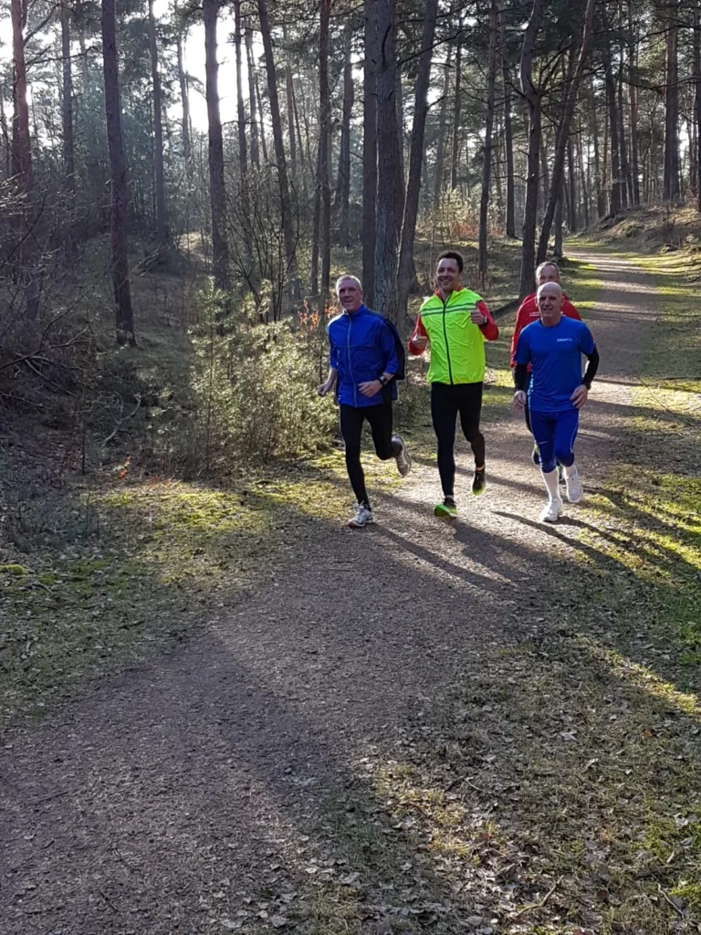 Vier Männer joggen auf einem Waldweg, umgeben von Bäumen und natürlicher Vegetation. Sie tragen sportliche Laufkleidung und scheinen motiviert und freundlich. Der sonnige Tag sorgt für eine angenehme Laufatmosphäre.