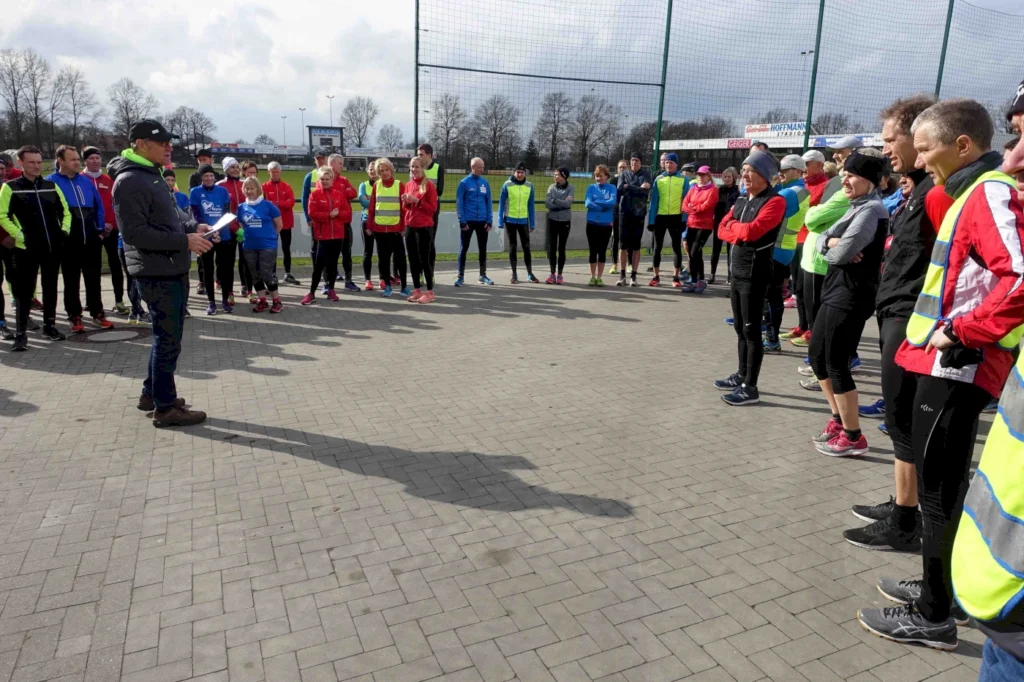 Gruppentraining im Freien mit Läufern, die einem Trainer zuhören, während sie sich auf eine Sportveranstaltung vorbereiten. Die Teilnehmer tragen verschiedene Laufbekleidungen und stehen auf einem gepflasterten Platz.