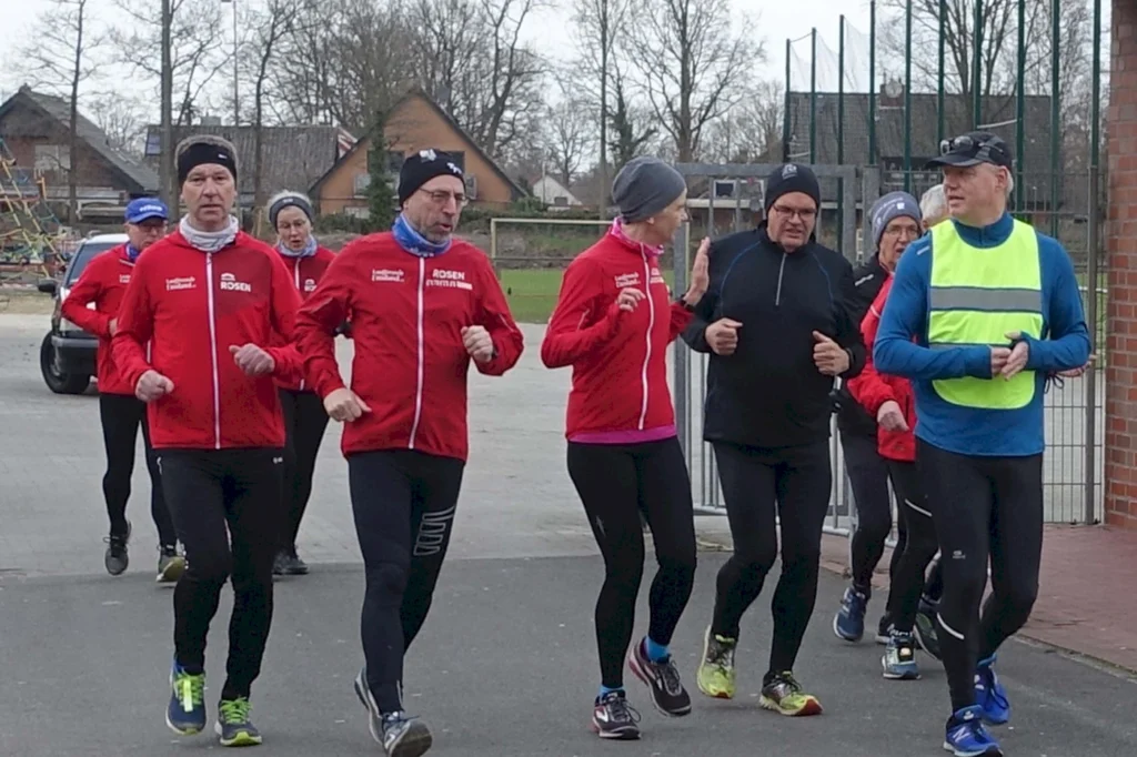 Gruppe von Läufern in roten Jacken, die gemeinsam im Freien joggen, umgeben von einer ländlichen Kulisse mit Bäumen und einem Sportplatz im Hintergrund.