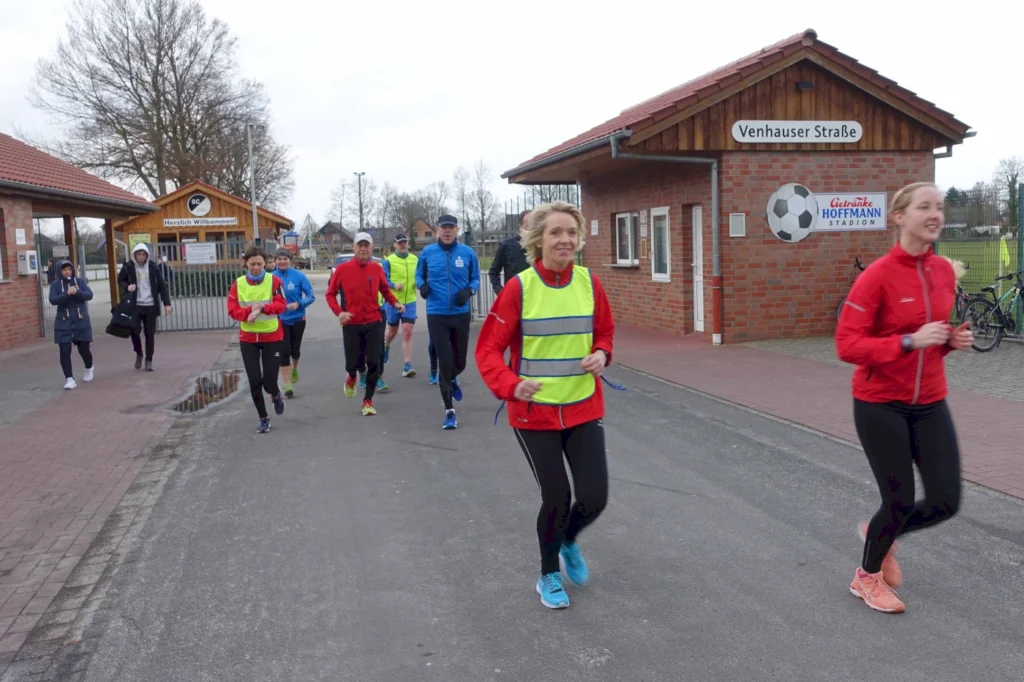 Läufer in bunten Sportoutfits joggen auf einer Straße vor einem Sportgebäude. Im Hintergrund ist das Schild "Herzlich Willkommen" sowie ein Fußballsymbol sichtbar. Die Szene vermittelt eine aktive, sportliche Atmosphäre.