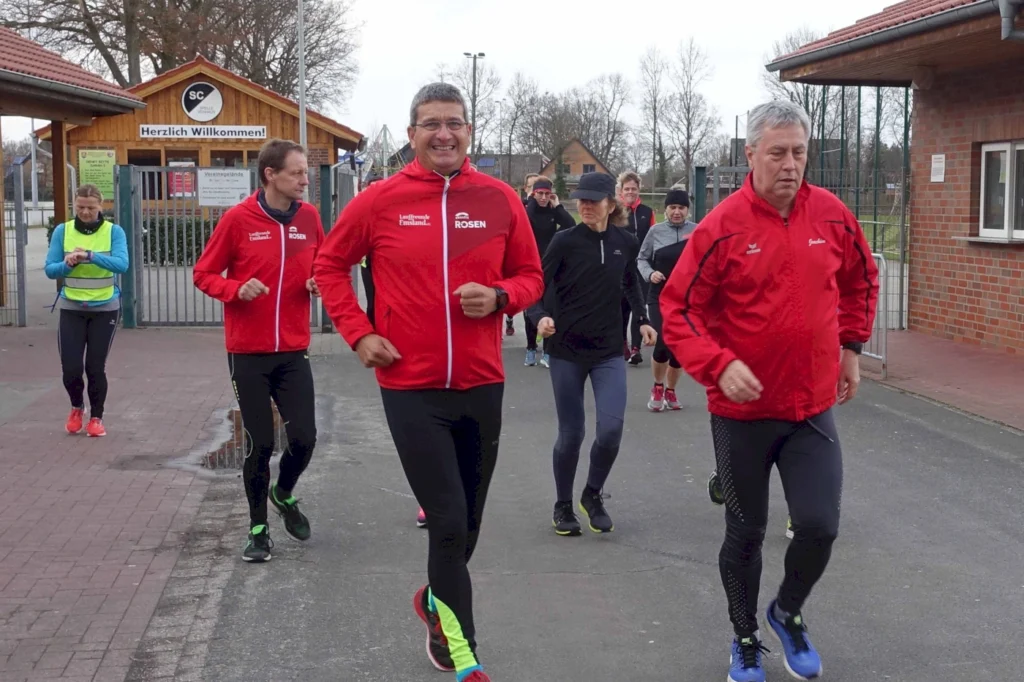 Gruppe von Läufern in roten Jacken, die fröhlich auf einem Sportgelände joggen, während sie an einem Vereinsgebäude vorbeigehen.