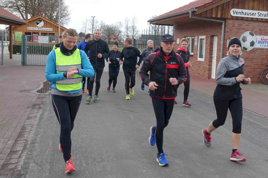 Läufergruppe in Sportkleidung joggt auf einer Straße vor einem Sportgebäude mit einem Willkommensschild. Eine Frau mit einer hellen Weste schaut auf ihre Uhr, während andere Teilnehmer in verschiedenen Outfits aktiv sind.
