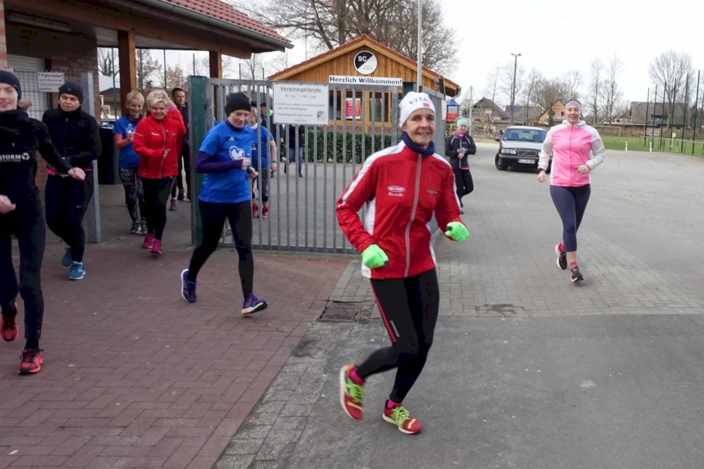 Läuferinnen und Läufer in Sportkleidung beim Training im Freien, vor einem Vereinsgebäude mit Willkommensschild. Die Gruppe besteht aus mehreren Personen, die fröhlich joggen, während es bewölkt ist.