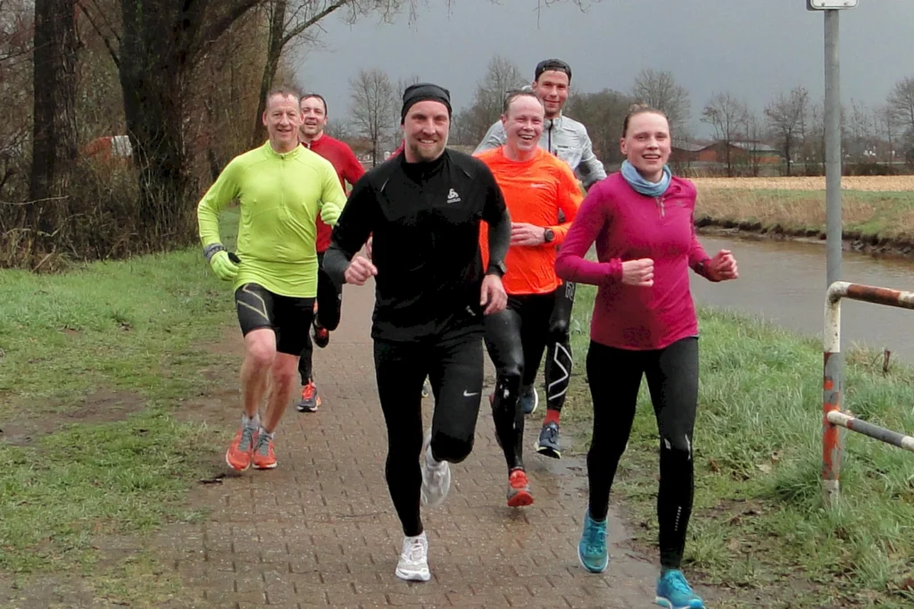 Gruppe von acht Läufern in sportlicher Kleidung, die auf einem befestigten Weg entlang eines Gewässers joggen, umgeben von grüner Vegetation und bewölktem Himmel.