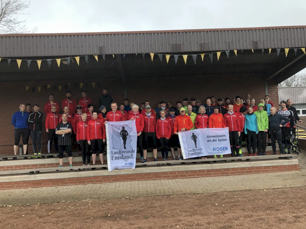 Gruppenfoto der Laufgemeinschaft Emsland, bestehend aus über 50 Mitgliedern in roten und bunten Laufjacken, die auf einer Tribüne stehen. Im Vordergrund wird ein Banner mit dem Slogan "Gemeinsam an die Spitze" präsentiert. Die Szene spiegelt den Teamgeist und die Begeisterung für das Laufen wider.