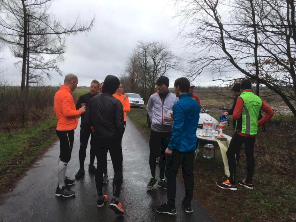 Gruppe von Läufern in leuchtenden Trainingsanzügen, die sich auf einem nassen Weg unterhalten, während ein Tisch mit Getränken und Snacks im Hintergrund steht. Bäume und ein bewölkter Himmel sind im Hintergrund sichtbar.