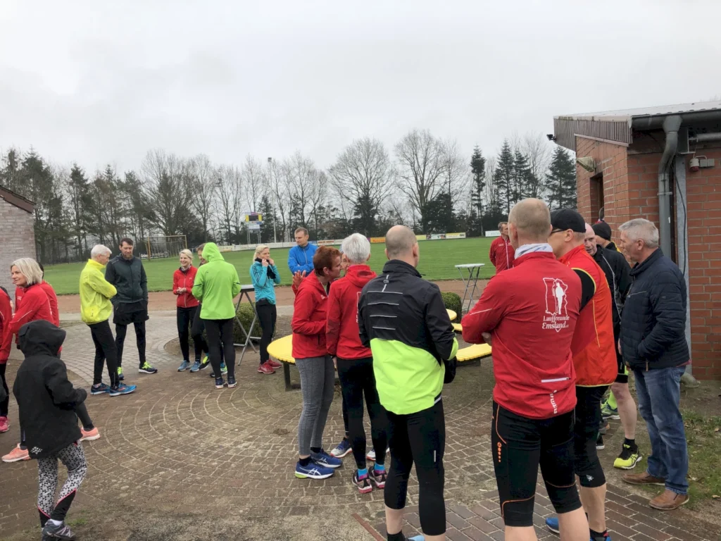 Gruppe von Läufern in Sportkleidung, die sich vor einem Vereinsgebäude versammeln, um sich auf ein Training oder einen Wettkampf vorzubereiten. Im Hintergrund ist ein Sportplatz mit Bäumen zu sehen.