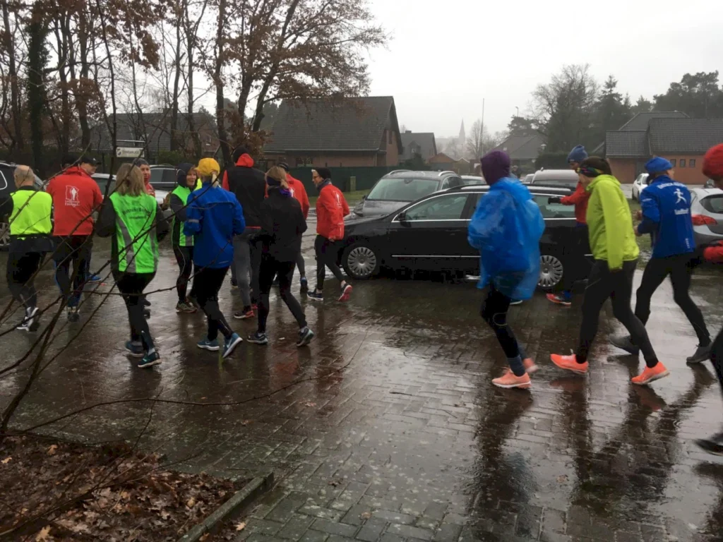 Läufer in bunten Jacken joggen bei Regen auf einem Parkplatz, umgeben von Bäumen und Wohnhäusern. Die Gruppe trägt Regenbekleidung und läuft trotz des schlechten Wetters.