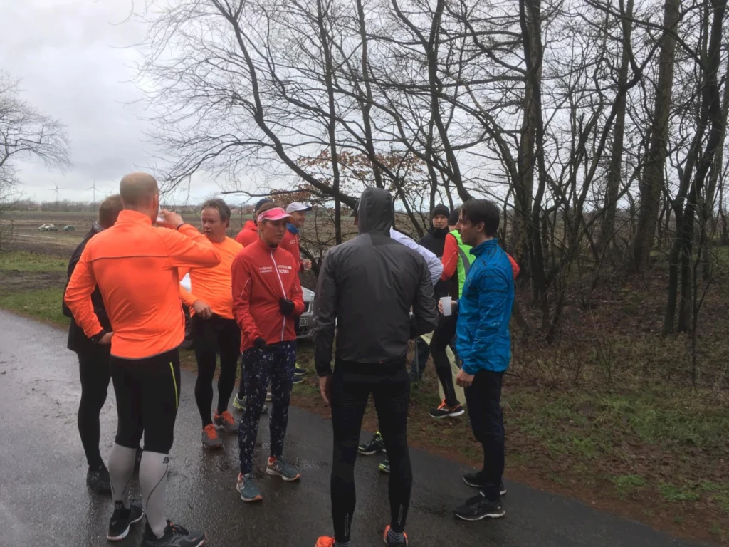 Gruppe von Läufern in leuchtenden Sportoutfits, die sich an einem regnerischen Tag auf einem Weg unter Bäumen versammeln, um sich vor einem Lauf auszutauschen und zu motivieren.
