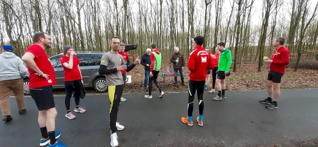 Läufergruppe in sportlicher Kleidung versammelt sich an einer Straße im Wald, einige halten Getränke in der Hand. Die Szene zeigt ein gemeinsames Event oder Training im Freien, umgeben von Bäumen und herbstlichem Laub.