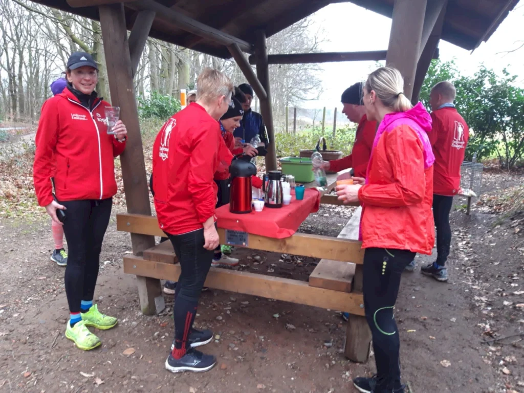 Gruppen von Läufern in roten Jacken stehen an einem Tisch im Freien, während sie Getränke und Snacks nach einem Lauf genießen. Im Hintergrund sind Bäume und ein Holzunterstand zu sehen.