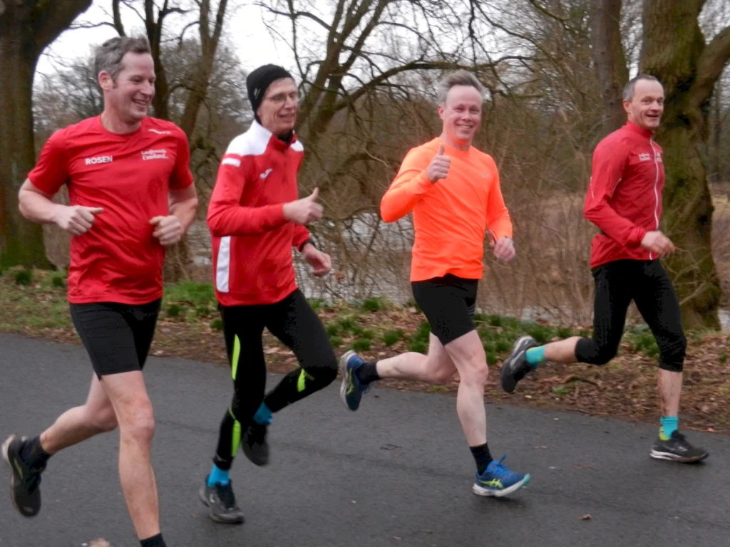 Vier Läufer in sportlicher Kleidung joggen auf einem Weg durch einen Park. Sie tragen rote und orange Shirts und zeigen eine positive Haltung. Im Hintergrund sind Bäume und eine ruhige Wasserfläche sichtbar.