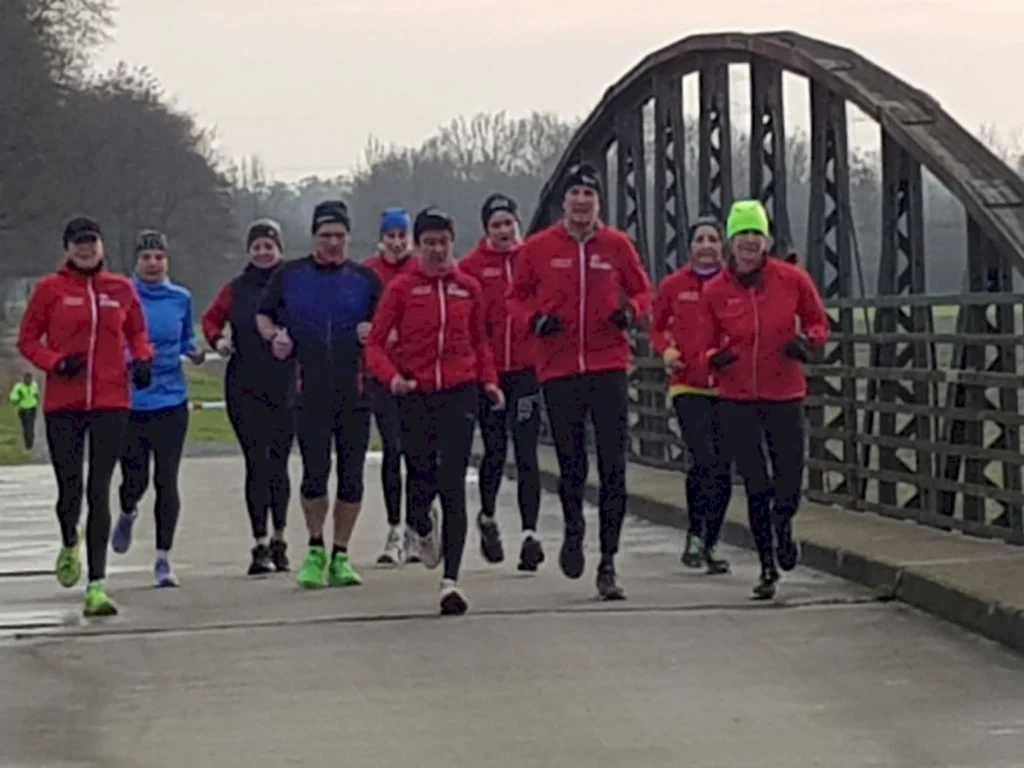 Gruppe von Läufern in roten Jacken, die auf einer Brücke joggen, bei kaltem Wetter und bewölktem Himmel, umgeben von einer grünen Landschaft.