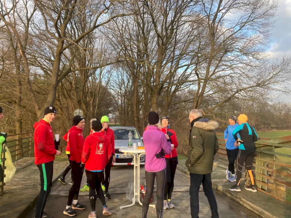 Gruppe von Läufern in sportlicher Kleidung, die auf einer Brücke stehen und sich unterhalten. Im Hintergrund sind Bäume und ein Auto zu sehen. Die Szene vermittelt eine freundliche und aktive Atmosphäre während einer Laufveranstaltung.