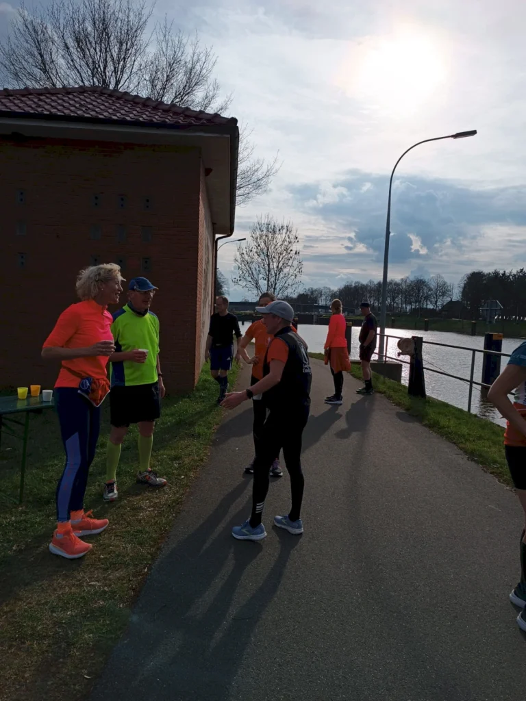 Gruppe von Läufern in bunten Sportoutfits unterhält sich an einem Laufstand am Ufer eines Gewässers, mit Bäumen im Hintergrund und bewölktem Himmel.