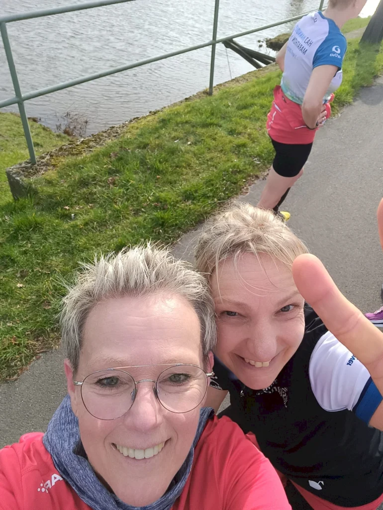 Zwei Frauen machen ein Selfie am Wasser entlang, während eine dritte Person im Hintergrund steht. Die Frauen tragen Sportkleidung und lächeln, während sie ihre Freizeitaktivitäten genießen. Grüne Wiese und ein Geländer sind im Bild sichtbar.