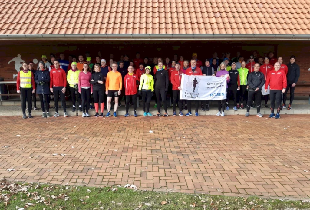 Gruppe von Läufern bei einem gemeinsamen Event der Laufgemeinschaft Emsland, versammelt unter einem Vordach, mit verschiedenen Laufoutfits und einem Banner, das die Gemeinschaft und den Zusammenhalt betont.