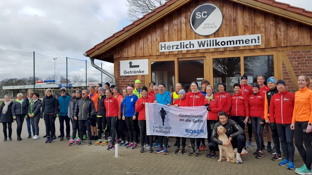 Gruppe von Läufern und Unterstützern vor dem SC Spelle-Venhaus, aufgenommen bei einer Veranstaltung für die Laufgemeinschaft Emsland. Die Teilnehmer tragen sportliche Kleidung und halten ein Banner mit dem Motto "Gemeinsam an die Spitze".