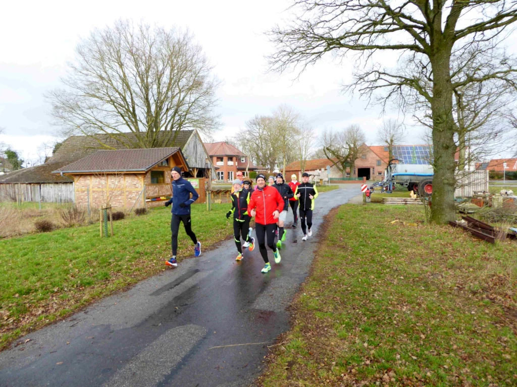 Gruppe von Läufern in Sportkleidung, die auf einer Landstraße in einer ländlichen Umgebung joggen, umgeben von Bäumen und landwirtschaftlichen Gebäuden. Ideal für Beiträge über Lauftraining, Outdoor-Aktivitäten oder ländliche Fitness.