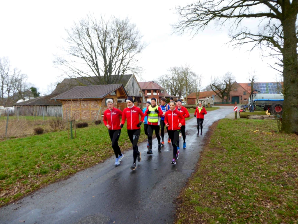 Eine Gruppe von Läufern in roten Jacken joggt auf einer ländlichen Straße, umgeben von Bäumen und Häusern. Die Teilnehmer scheinen an einem Lauftraining oder einer Veranstaltung teilzunehmen, während es bewölkt ist.