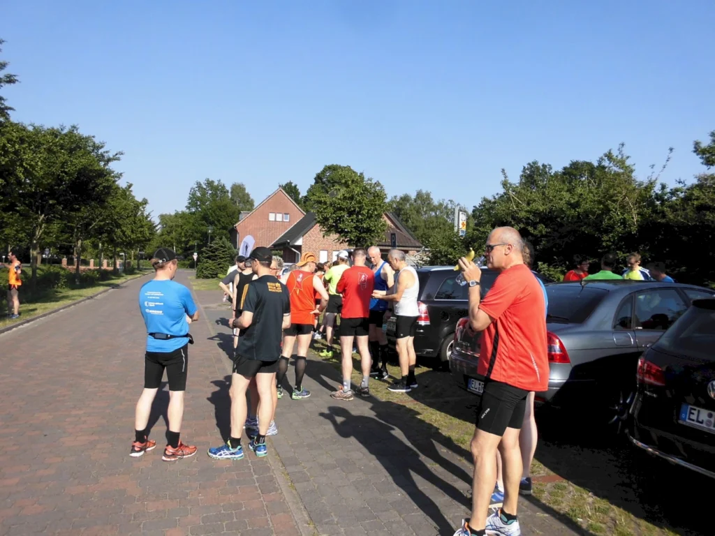 Läufer in Sportkleidung versammeln sich auf einer Straße vor einem Veranstaltungsort, um an einem Wettlauf teilzunehmen. Die Szenerie zeigt eine sonnige Atmosphäre mit grünen Bäumen und geparkten Autos.