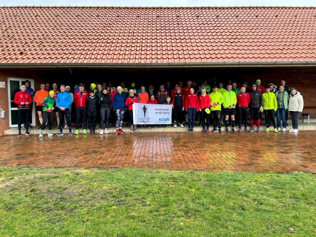 Gruppenfoto von Läufern in bunter Sportkleidung, die sich bei einem Lauf-Event unter einem Dach versammeln. Im Hintergrund ist ein Banner mit der Aufschrift „Gemeinsam an die Spitze“ zu sehen. Das Wetter ist regnerisch, und die Teilnehmer lächeln und posieren für das Foto.